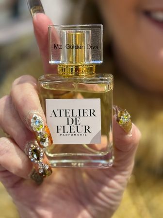 Close-up of a glass perfume bottle with a gold cap and white label held by a hand showcasing ornate rhinestone and floral nail art — beauty salon style.