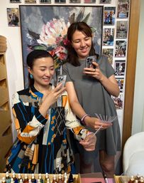 Two smiling women in a beauty boutique taking selfies and comparing nail-color swatch fans over a counter of small dropper bottles, floral painting and photo collage on the wall behind them.