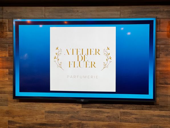 Wall-mounted flat-screen on a wood-paneled wall displaying a blue-framed digital sign with a white square, gold floral motifs and elegant gold lettering advertising a parfumerie.