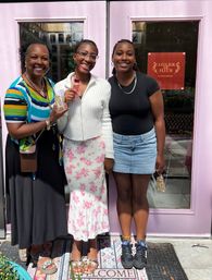 Three smiling women pose in front of a pink storefront door on a sunny city sidewalk, holding perfume sample bottles — one in a colorful striped top, one in a white sweater with a floral midi skirt, and one in a black tee with a denim mini skirt.