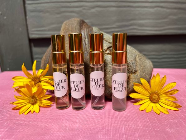 Four glass travel perfume roller bottles with shiny gold caps arranged on a pink textured cloth between bright yellow daisy flowers, set against smooth river rocks and gray wooden siding backdrop.