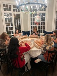People gathered around a cozy home dining table for a scent‑blending party, dozens of small perfume and essential oil bottles on trays, chandelier and a "Happy Birthday" balloon overhead.
