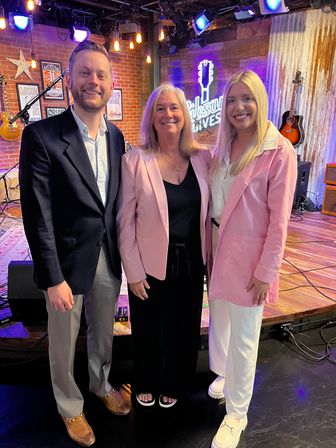 Three smiling adults posing on a cozy music studio stage with exposed brick, hanging Edison bulbs, guitars and a neon guitar sign; two women in pink blazers and a man in a dark blazer.
