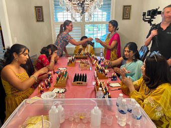 Women in colorful traditional dresses seated around a dining table under a crystal chandelier, sampling and mixing dozens of small dropper bottles and vials in a bright home workshop setting.
