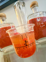 Close-up of a bright pink sparkling drink in a plastic cup topped with red slush, staged in front of glass jars and wooden-stopper bottles on a café counter.