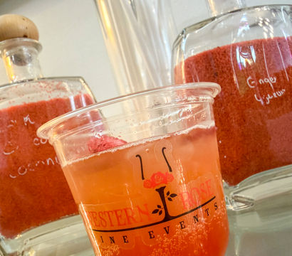 Close-up of a bright pink sparkling drink in a plastic cup topped with red slush, staged in front of glass jars and wooden-stopper bottles on a café counter.