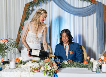 Bride in lace gown pours a giant champagne bottle for her seated partner at an indoor wedding reception table adorned with orange and white roses, candles, greenery and a blue-draped wooden arch