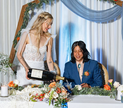 Bride in lace gown pours a giant champagne bottle for her seated partner at an indoor wedding reception table adorned with orange and white roses, candles, greenery and a blue-draped wooden arch