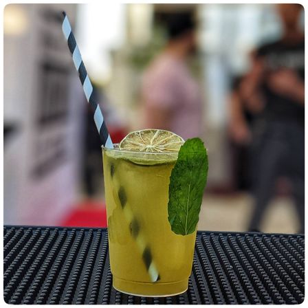 Refreshing iced lime-mint cocktail in a clear cup with black-and-white striped straw, dried lime wheel and fresh mint leaf on a bar mat against a blurred outdoor crowd.