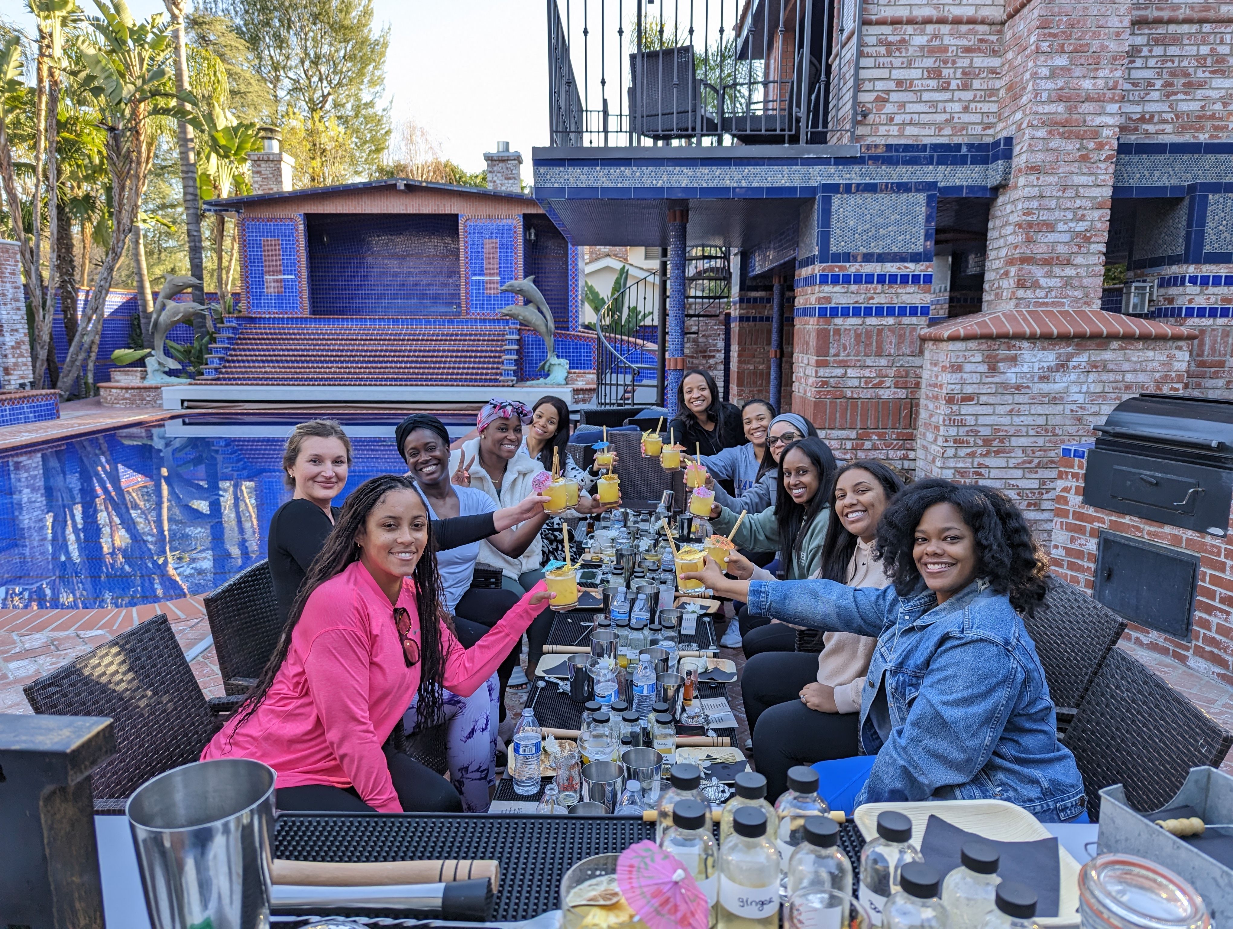Group of women toasting with yellow cocktails at a long table on a brick backyard patio beside a blue-tiled pool and dolphin statues — festive poolside brunch celebration.