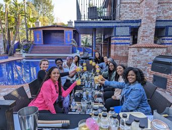Group of women toasting with yellow cocktails at a long table on a brick backyard patio beside a blue-tiled pool and dolphin statues — festive poolside brunch celebration.