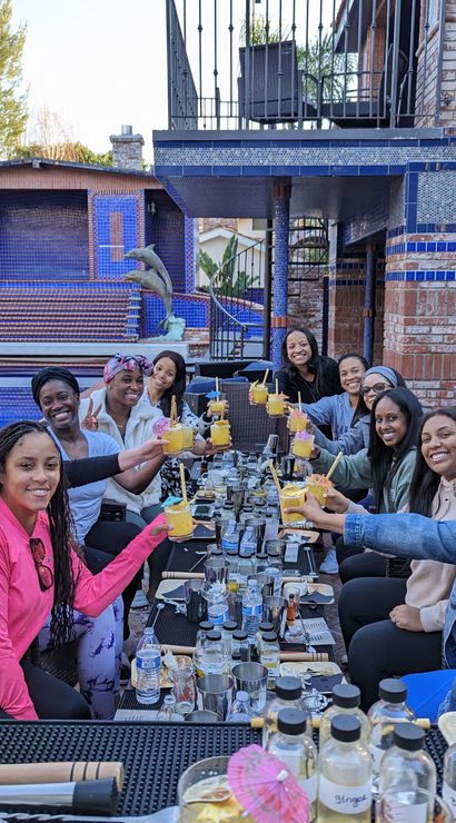 Group of women toasting with yellow cocktails at a long table on a brick backyard patio beside a blue-tiled pool and dolphin statues — festive poolside brunch celebration.