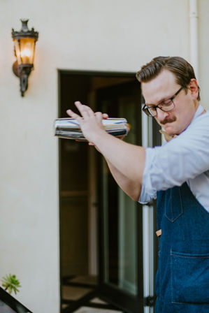 Bartender in denim apron shaking a stainless steel cocktail shaker on a sunlit patio beside a vintage wall lantern and open doorway