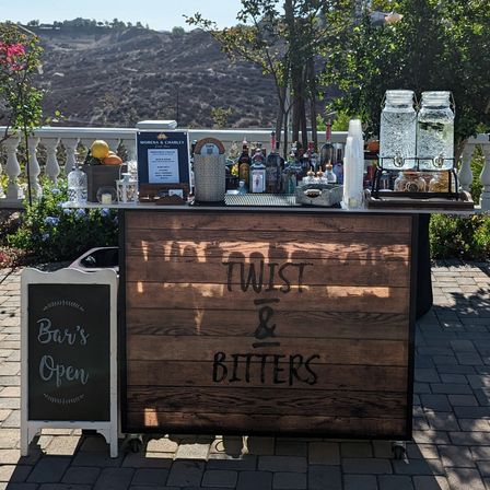 Sunny outdoor mobile cocktail bar on a patio with a rustic wooden front panel, rows of liquor bottles, glass beverage dispensers with cups, fresh citrus and bar tools, a small chalkboard sign, and rolling hills in the background.