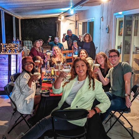 Cheerful group of friends toasting with margaritas around a long table on a covered backyard patio at night, colorful DIY bar, string lights and festive vibe.