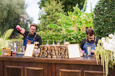 Outdoor garden bar with two bartenders in denim aprons and bow ties mixing cocktails behind a rustic wooden counter topped with large 'BAR' letters, cocktail tools, flowers and lush tropical greenery