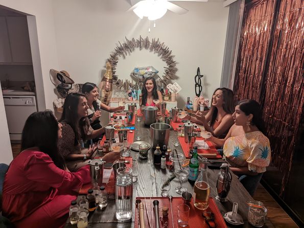 Friends at an apartment cocktail-making party around a long dining table, laughing and mixing drinks with shakers, bottles, glassware and garnishes, framed by a metallic fringe curtain and a driftwood wreath for a festive night in.