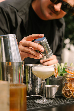 Bartender adding a zesty finishing touch by sprinkling citrus over a frothy craft cocktail in a coupe glass with dried orange garnish at an outdoor bar.