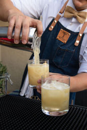 Bartender pouring a frothy yellow cocktail from a metal shaker into a rocks glass with ice, wearing a denim apron and bow tie
