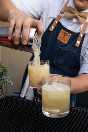 Bartender pouring a frothy yellow cocktail from a metal shaker into a rocks glass with ice, wearing a denim apron and bow tie