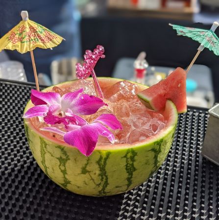 Half watermelon used as a cocktail bowl filled with crushed ice, garnished with a pink orchid, watermelon wedge, colorful paper umbrellas and a sparkly stirrer on a bar mat — tropical watermelon cocktail.