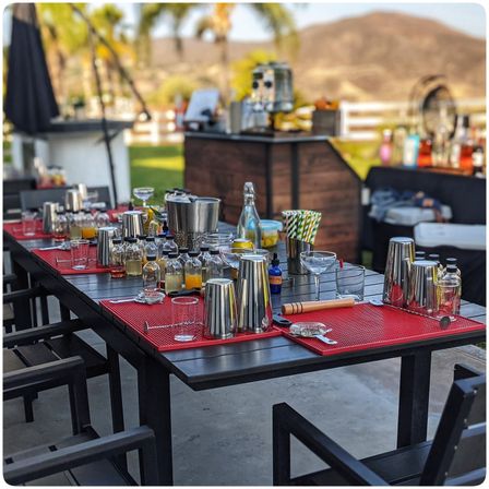 Backyard patio cocktail station with stainless shakers, mixers, glassware and striped straws on red bar mats, palm-tree and hillside backdrop