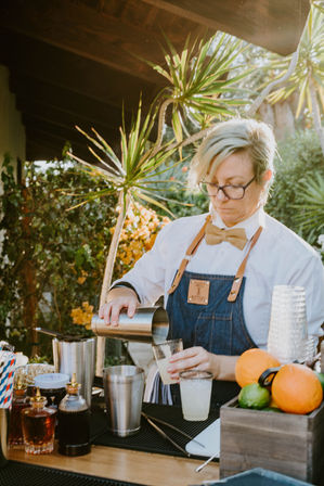 Outdoor bartender in a denim apron and bow tie pouring a shaken citrus cocktail into a glass at a sunny backyard garden bar with shakers, bitters bottles and fresh oranges nearby.
