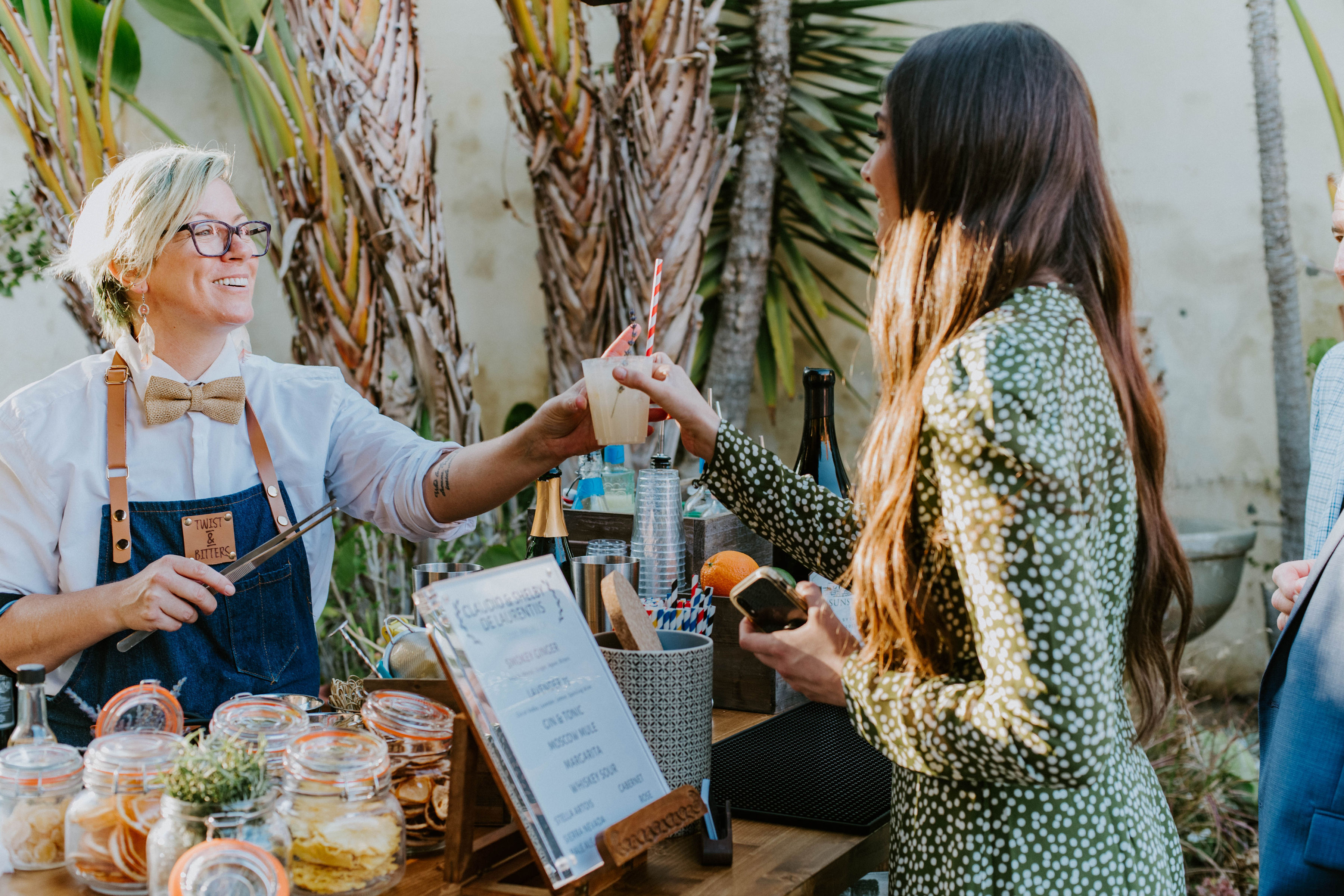 Outdoor garden bar scene as a smiling vendor in an apron and bow tie hands a craft cocktail with a striped straw to a guest in a green patterned dress, jars of garnishes and a menu on the wooden counter.