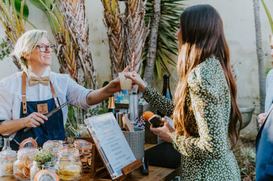 Outdoor garden bar scene as a smiling vendor in an apron and bow tie hands a craft cocktail with a striped straw to a guest in a green patterned dress, jars of garnishes and a menu on the wooden counter.