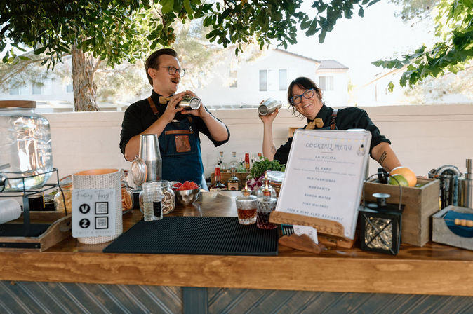Two smiling bartenders shaking cocktail shakers behind a wooden outdoor cocktail bar under leafy trees, surrounded by jars, fruit, bottles and a visible cocktail menu in a sunlit suburban backyard.