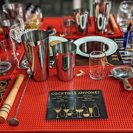 Close-up of a cocktail bar mixology station on a red rubber mat with polished stainless steel shakers, jigger, mixing spoon, strainer, muddler, measuring glass and a “Cocktails Anyone?” menu card, ready for happy hour.