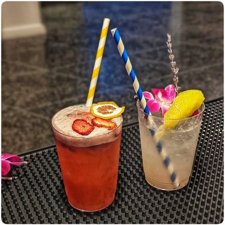 Two colorful summer cocktails on a black bar mat: a frothy strawberry-red drink topped with dried citrus slices and a yellow-striped paper straw, and a sparkling lemon soda garnished with a lemon wedge, pink orchid and lavender sprig with a blue-striped paper straw.