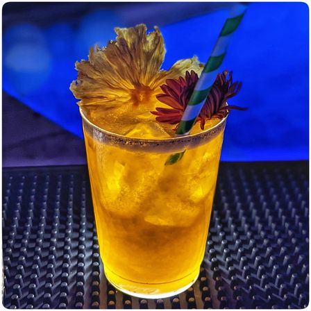 Neon-lit tropical cocktail on a bar mat — bright yellow crushed-ice drink garnished with dried pineapple, red flower and green-striped paper straw.