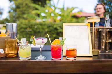 Four colorful cocktails lined up on a wooden outdoor bar — a green citrus drink with a peppered rim, a creamy floral coupe garnished with leaves and petals, a bright red cocktail with an orange slice and decorative pick, and an amber whiskey — blurred menu frame and greenery in the background.