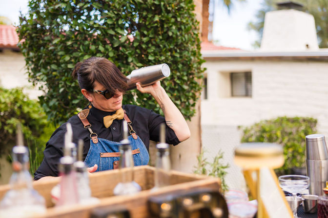 Bartender in denim apron and bow tie shaking a metal cocktail shaker at an outdoor garden bar on a sunny patio, bottles and glassware in the foreground.