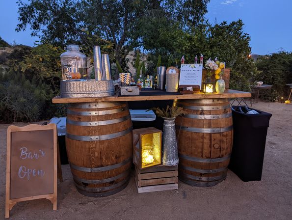 Rustic outdoor bar at dusk with wooden wine barrels as a counter, galvanized drink dispenser and metal cups, illuminated lanterns, a drink menu and chalkboard sign reading "Bar's Open" in a garden setting
