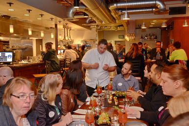 Chef speaking to diners at a long table in a busy modern restaurant with open kitchen, exposed ductwork, pendant lights, cocktails and shared appetizers.