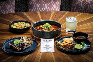 Restaurant table spread: Mexican‑inspired stew in a bowl, cheesy quesadillas with lime and dipping broth, braised meat with slaw, seasoned rice and a cocktail.