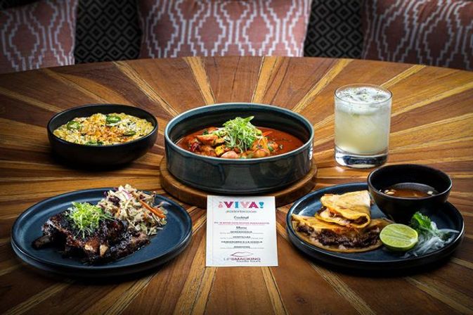 Restaurant table spread: Mexican‑inspired stew in a bowl, cheesy quesadillas with lime and dipping broth, braised meat with slaw, seasoned rice and a cocktail.