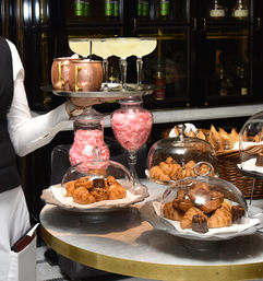 Server holding a tray of cocktails and copper mugs above a marble cafe table filled with croissants, canelés and assorted pastries under glass cloches, with jars of pink rose petals — elegant bakery and cocktail display.
