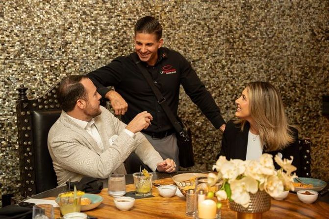 Two diners seated at a candlelit restaurant table smile and chat with a standing server against a gold mosaic wall, with drinks, small plates, and an orchid centerpiece on the wooden table.
