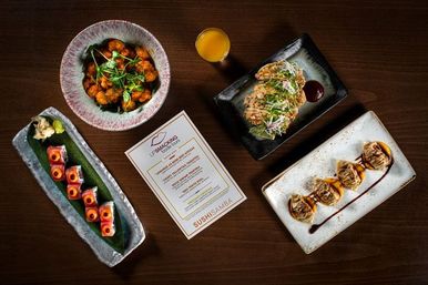 Top-down restaurant shot of colorful sushi rolls, dumplings, saucy fried bites, a small glass of juice and a menu arranged on a dark wood table — Japanese-fusion spread.
