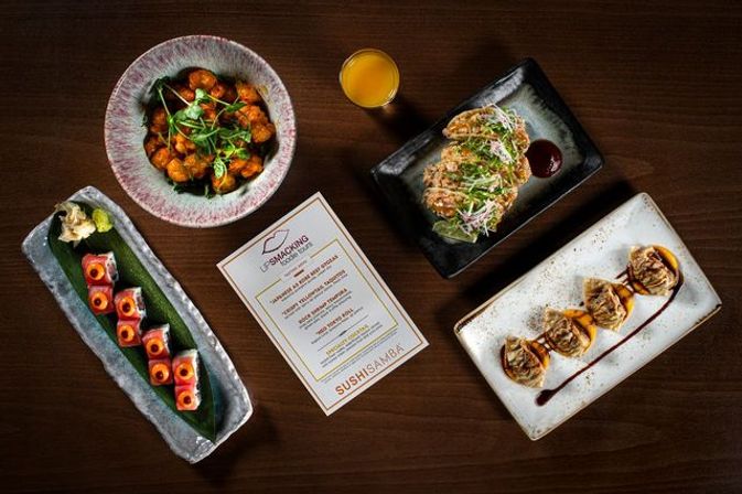 Top-down restaurant shot of colorful sushi rolls, dumplings, saucy fried bites, a small glass of juice and a menu arranged on a dark wood table — Japanese-fusion spread.