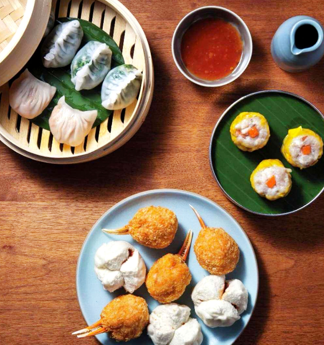 Top-down view of a Chinese dim sum spread on a wooden table: bamboo steamer with translucent and green dumplings, a plate of fried crab-claw croquettes and steamed BBQ pork buns, siu mai on a banana leaf, and a bowl of chili dipping sauce.