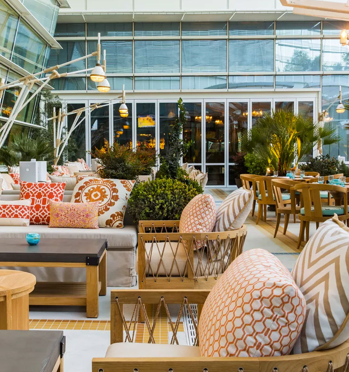 Vibrant urban rooftop patio lounge with wooden sofas and chairs, orange patterned cushions, potted greenery and modern hanging lamps outside a glass-fronted restaurant