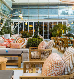 Vibrant urban rooftop patio lounge with wooden sofas and chairs, orange patterned cushions, potted greenery and modern hanging lamps outside a glass-fronted restaurant