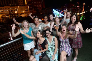Energetic group of friends cheering with drinks on a rooftop terrace at night, city lights and neon signs glowing in the background — urban nightlife scene.