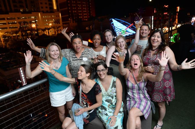 Energetic group of friends cheering with drinks on a rooftop terrace at night, city lights and neon signs glowing in the background — urban nightlife scene.
