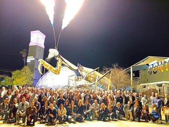 Large group photo at night in a downtown outdoor plaza in front of a giant metal praying mantis public art installation shooting flames from its antennae.