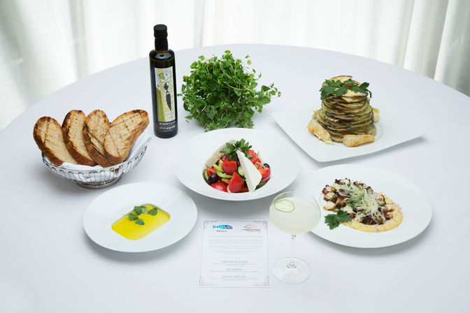 Mediterranean-inspired spread on a white tablecloth: toasted artisan bread in a basket, bottle of olive oil and fresh herbs, Greek salad with feta and olives, stacked roasted eggplant tower, octopus on corn puree, olive oil dipping plate, and a cucumber-garnished cocktail.
